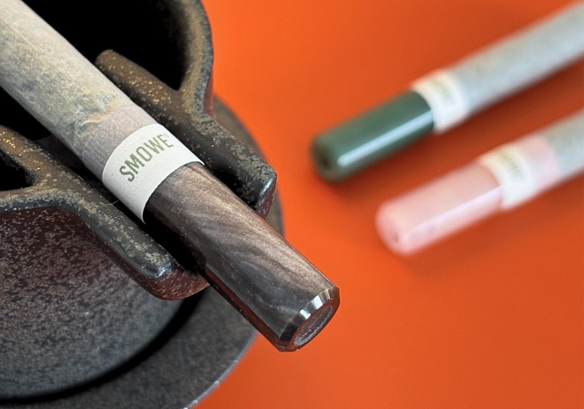 SMOWE J-Band Papierbanderole an Kräuter-Zigarette mit dunklem Glas-Tip auf Halterung.