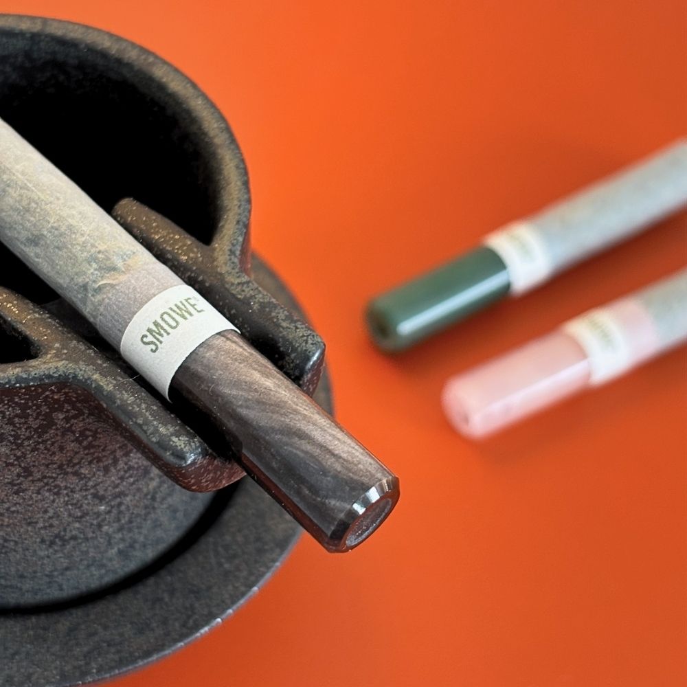 Eingedrehter Joint mit dunklem SMOWE Glasfilter und weißer Markenbanderole in einer Keramikschale auf orangefarbenem Untergrund.